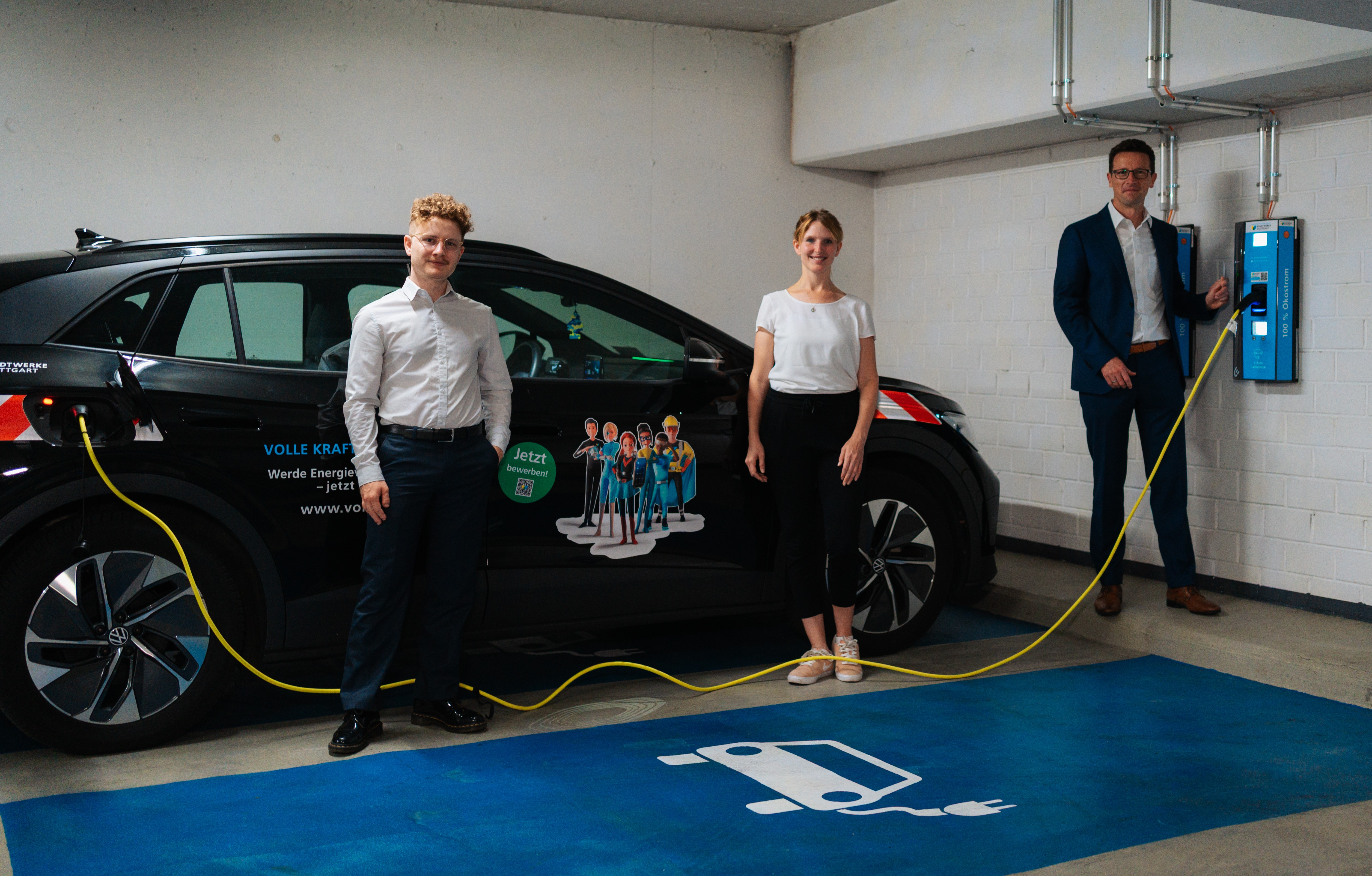 Drei Personen stehen neben einem schwarzen E-Auto in einer Garage. Das Auto lädt an einer blauen Ladesäule mit gelbem Kabel.