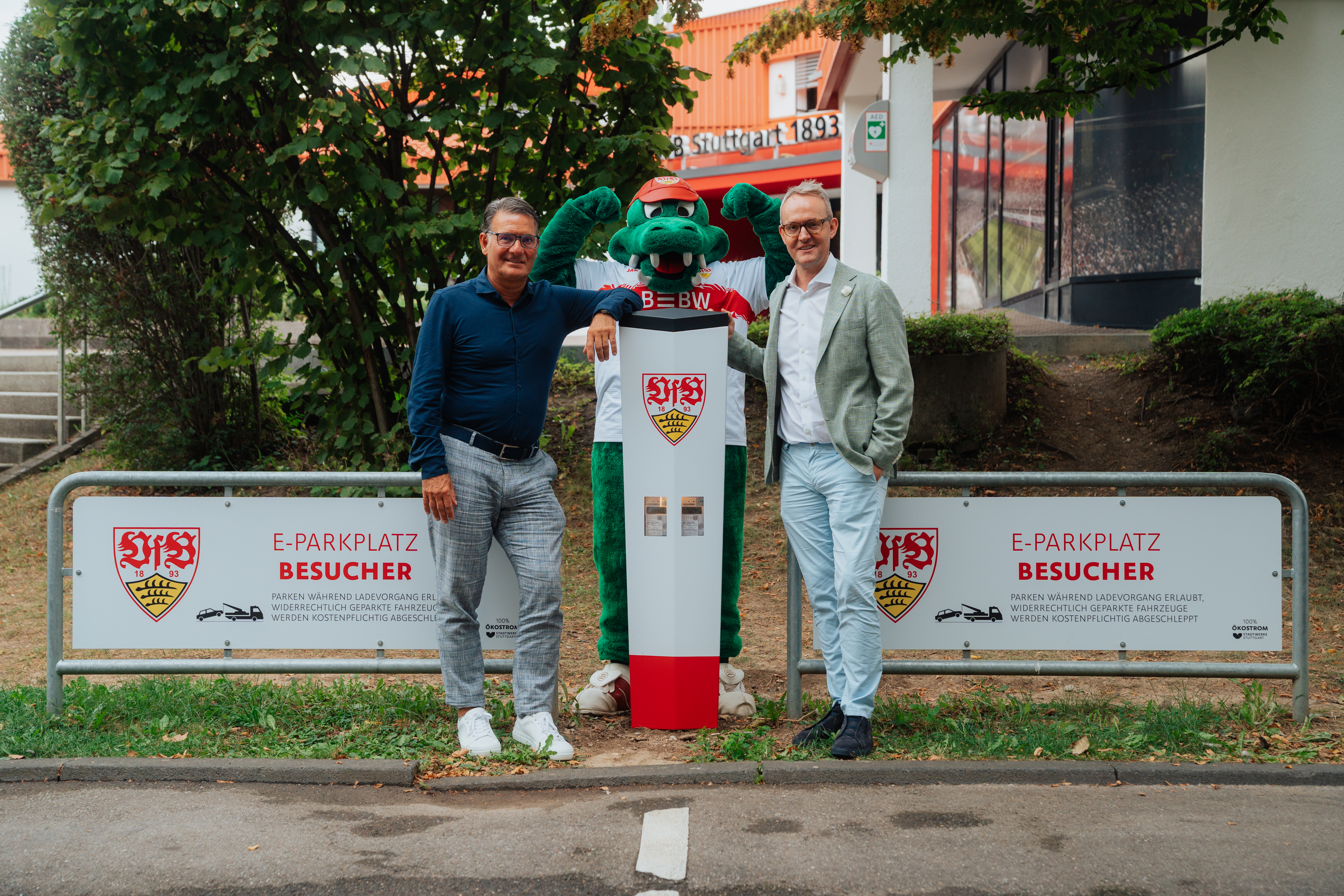 Zwei Männer und ein grünes Maskottchen stehen vor einer E-Ladesäule mit VfB-Logo auf einem Besucherparkplatz.